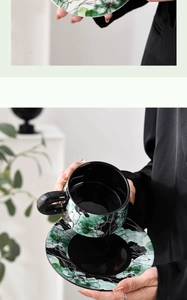 Close-up of hand holding black ceramic coffee cup with green floral patterns and saucer