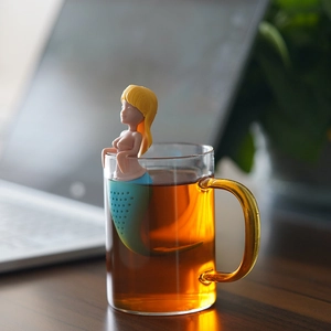 Whimsical mermaid silicone tea infuser hanging on the edge of a clear glass cup