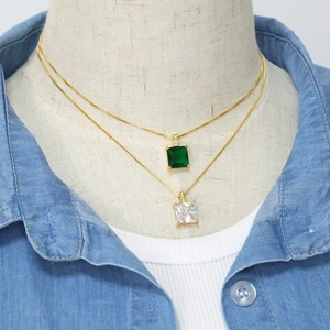Détail de colliers dorés superposés avec pendentifs carrés en zircon vert et transparent