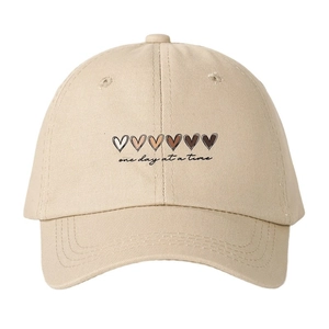 Beige baseball cap with gradient heart icons and one day at a time script embroidery