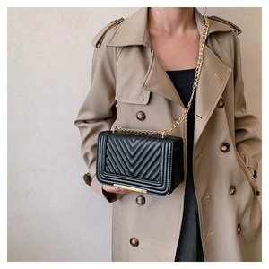Detail of black V-quilted crossbody bag with gold chain strap on a trench coat
