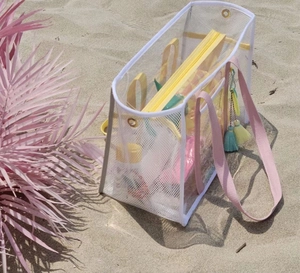 Detailed view of a transparent mesh beach bag with pink straps and tassel on sandy beach
