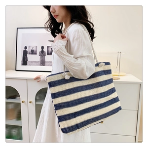 Blue and beige striped straw tote bag with rope handles worn by a model in a cozy room