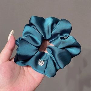 Hand holding a large teal satin scrunchie with silky folds and a silver metal tag
