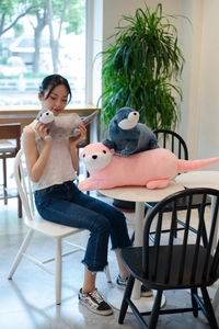 Plush otter dolls in grey, blue, and pink showing three sizes in a cozy cafe setting