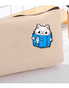 Close-up of white cat enamel pin reading a blue book on a beige canvas backpack