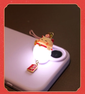 Close-up of a glowing white Maneki Neko lucky cat charm with a red Ping'an tag on phone