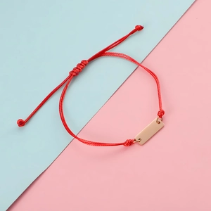 Detail of red braided cord bracelet with polished gold rectangle bar on pastel background