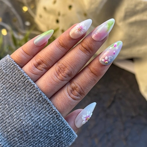 Detail of almond press-on nails with 3D white flowers, gold beads, and pink gradient finish