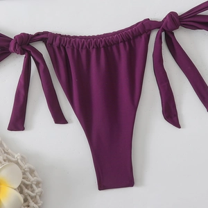 Dettaglio dello slip bikini viola scuro con laccetti laterali e arricciature su sfondo spiaggia