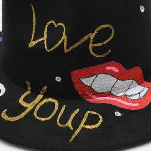 Detail of black felt fedora featuring hand-painted gold lettering and a punk lip graffiti