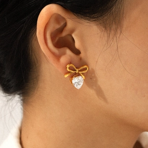 Close-up of golden bow stud earring with heart-shaped zircon drop on a female ear
