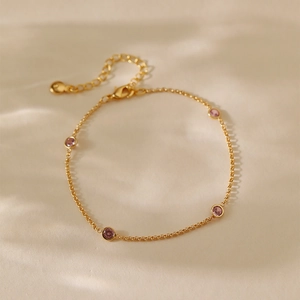 Elegante pulsera de cadena dorada con circonitas rosas redondas sobre tela beige y luz cálida