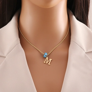 Detail of gold M-letter pendant and blue evil eye bead on a fine stainless steel chain