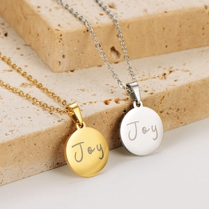 Gold and silver stainless steel Joy round pendant necklaces on a beige stone base