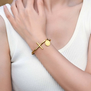 Detail of a gold stainless steel open bangle with cross and oval medal pendant on a wrist