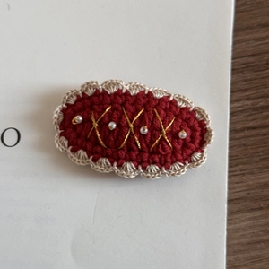 Detail of crochet hair clip featuring red yarn, pearl accents, and gold thread patterns