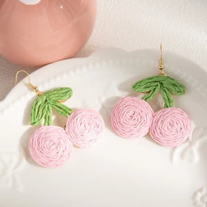 Close-up of handmade woven pink cherry earrings with green leaves on a white ceramic plate