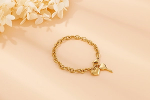 Detailed view of gold stainless steel bracelet with heart and key charms on peach background