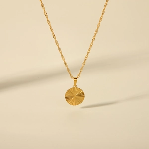 Detail of a golden necklace featuring a circular sunburst textured pendant on beige back