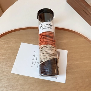 Three earth-toned braided hair ties in a clear cylindrical box with a minimalist label