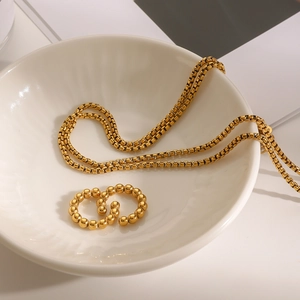 Close-up of a gold square chain necklace and beaded ear cuffs in a white ceramic bowl