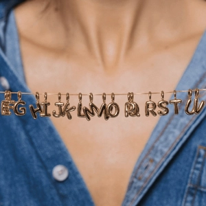 Close-up of gold-plated balloon letter pendants on a model wearing a blue denim shirt