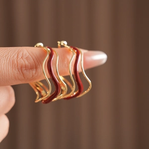 Close-up of wavy gold hoop earrings with red enamel coating held by fingers