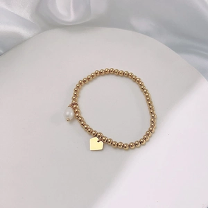 Close-up of golden beaded stretch bracelet with pearl charm and heart tag on white silk
