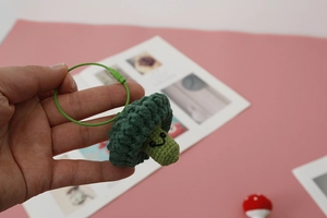 Close-up of handmade crochet broccoli keychain with green wire loop on pink desk