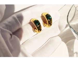Macro shot of gold-plated C-shaped earrings featuring emerald green oval zircon stones
