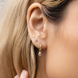 Detail of model wearing a gold heart stud, purple gem earring and gold hoop drop set