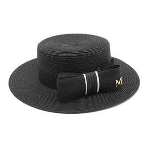 Detail of a black woven boater hat with a bow and gold M letter metal accessory
