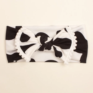 Soft baby headwrap featuring cow print with a large bow and white pom-pom trim detail