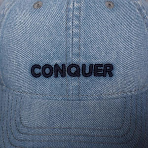 Close-up of CONQUER blue embroidery on light blue washed denim baseball cap texture