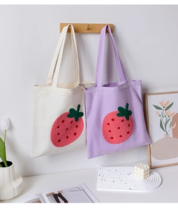 Detail of embroidered strawberry tote bags in beige and purple on a wooden hook