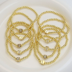 18k gold beaded bracelets with sparkling cubic initial charms displayed in a ceramic bowl