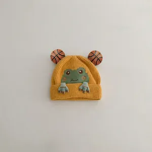 Detail of yellow knit baby beanie featuring a cute green cartoon frog with eye pom-poms