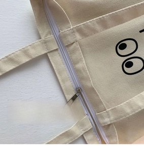 Primer plano del bolso de lona beige con cierre de cremallera y diseño de ojos grabado