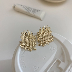 Detailed close-up of golden irregular square earrings with woven texture on a white lid