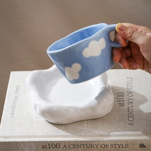 Detail of blue ceramic cloud mug held by hand with white cloud-shaped saucer on a magazine