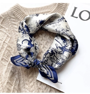 Detail of a silk neck scarf with blue forest print knotted on a beige knit sweater