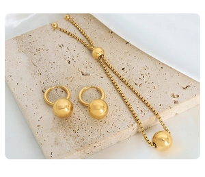 Detail of gold-plated steel jewelry set featuring ball pendants on beige stone background