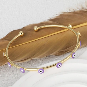 Golden evil eye open bangle with purple enamel details on a textured white display stand