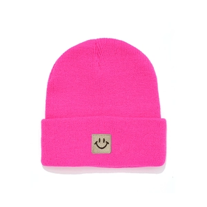 Detail of fuchsia knit beanie featuring a square black smiley face patch and ribbed texture