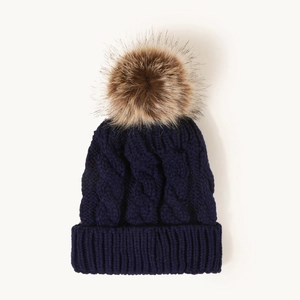 Detail of navy cable knit beanie with a fluffy brown faux fur pom pom and thick ribbed cuff