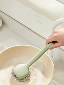 Green long-handle kitchen brush with steel wool scrubbing a soapy pan in a sink