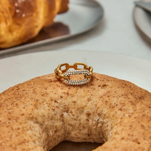 Anello a catena dorata in titanio con zirconi ovali su sfondo di pasticceria di lusso