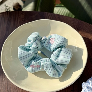 Detailed view of a light blue striped scrunchie with pink ACED embroidery on a beige plate