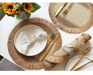 Top view of woven round placemats with fringe edge, napkin rings and gold cutlery on white desk